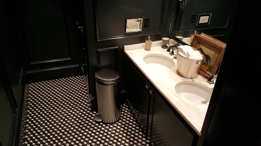 Modern bathroom with dark walls, patterned floor, double sink vanity, and a trash can.
