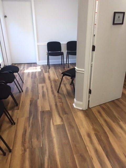 Empty hallway with black chairs lining the walls and a white door open on the right, wood-patterned floor.