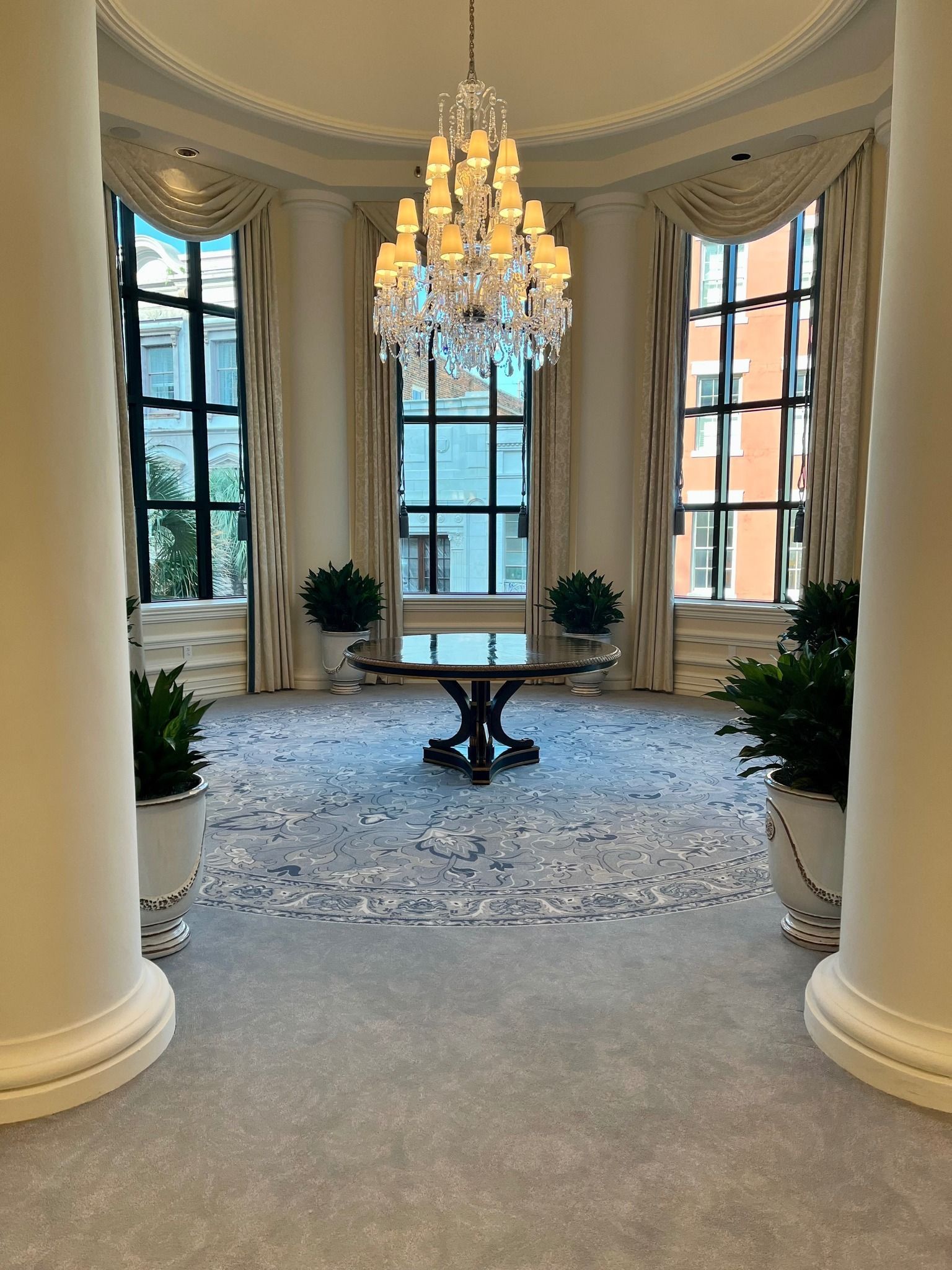Elegant foyer with chandelier, round table, large windows, and potted plants