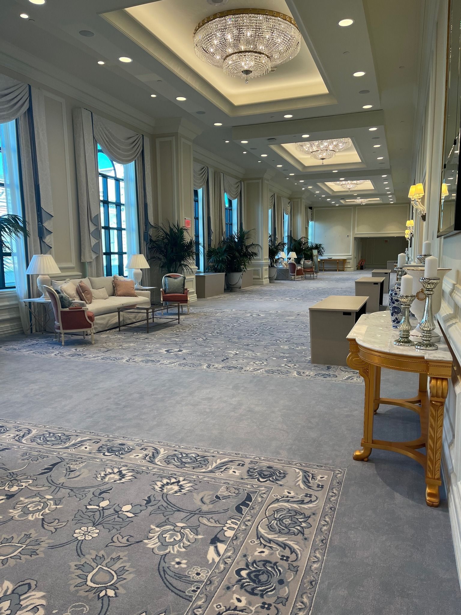 Elegant hotel ballroom with patterned carpet, chandeliers, tall windows, and seating along the walls
