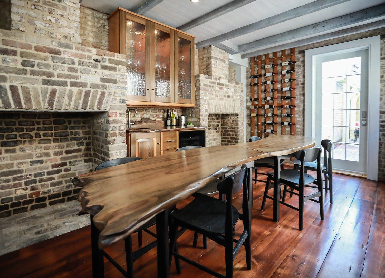 Rustic dining room with long wooden table, brick fireplace, exposed beams, and glass door opening outside