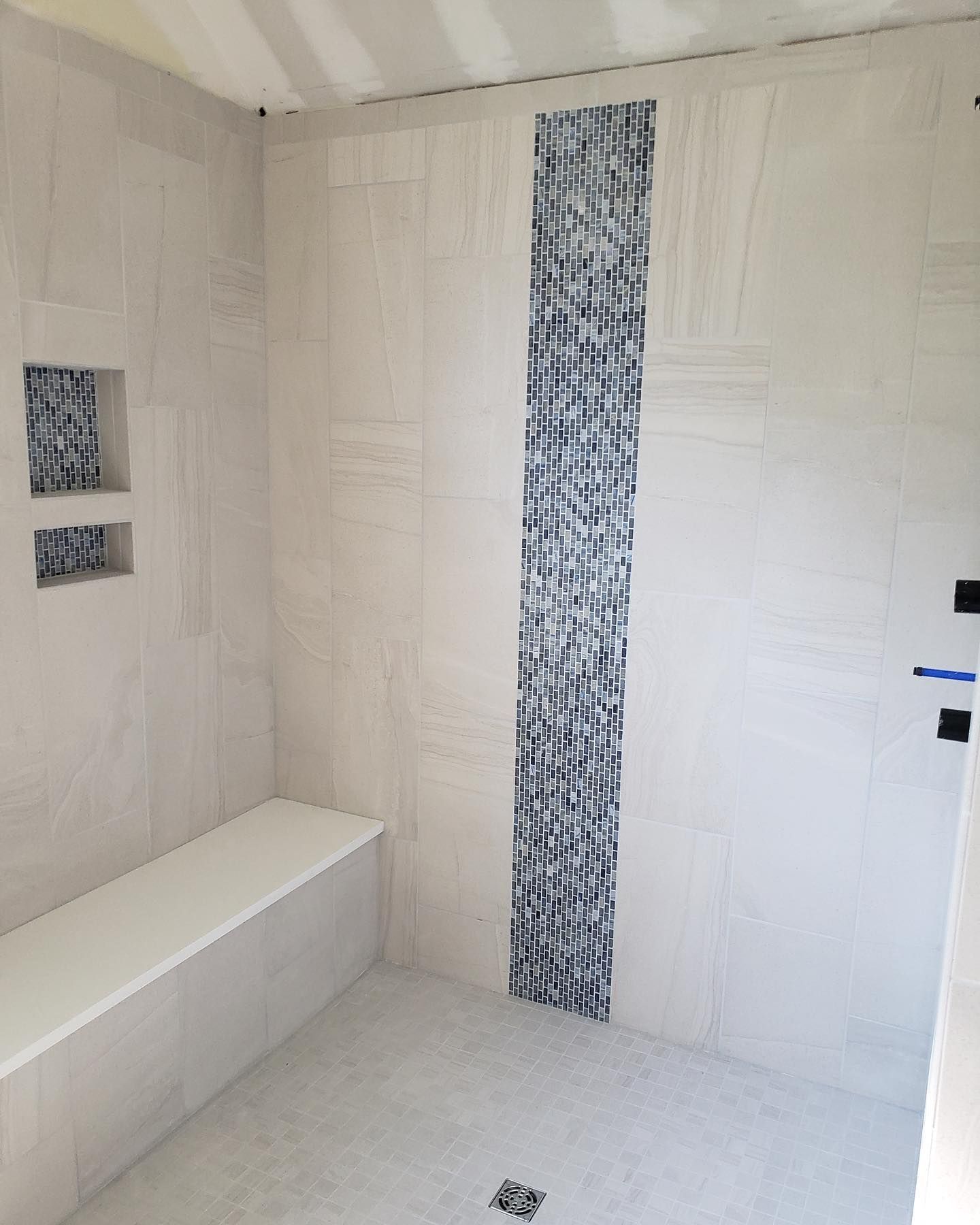 Modern bathroom with white tiled walls, a vertical blue-gray mosaic strip, bench, and shower floor drain