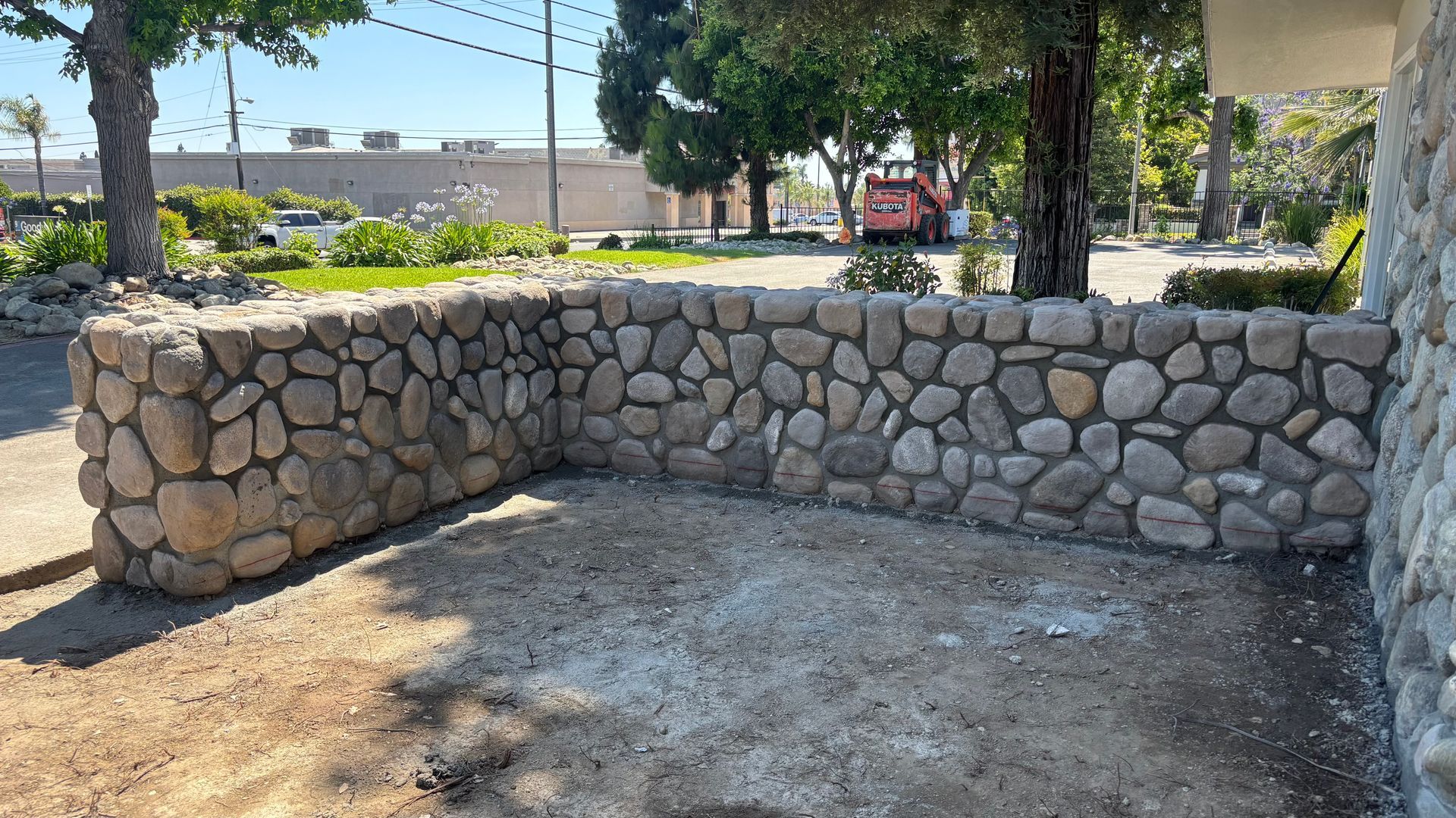 Stone-walled outdoor corner with a gravel floor beside a sidewalk and trees