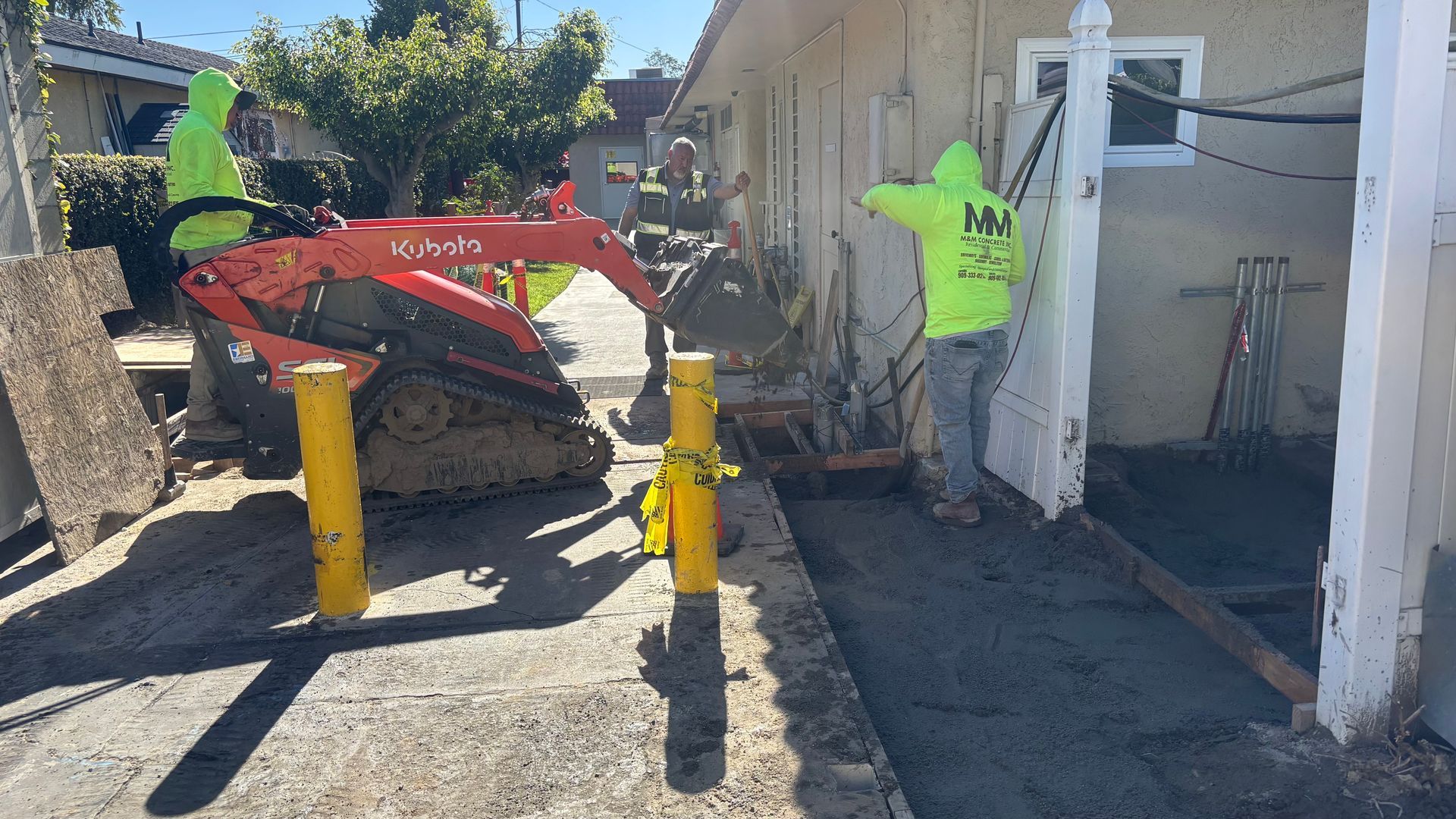 Worker beside red skid steer in a narrow alley, near yellow bollards and a white building.