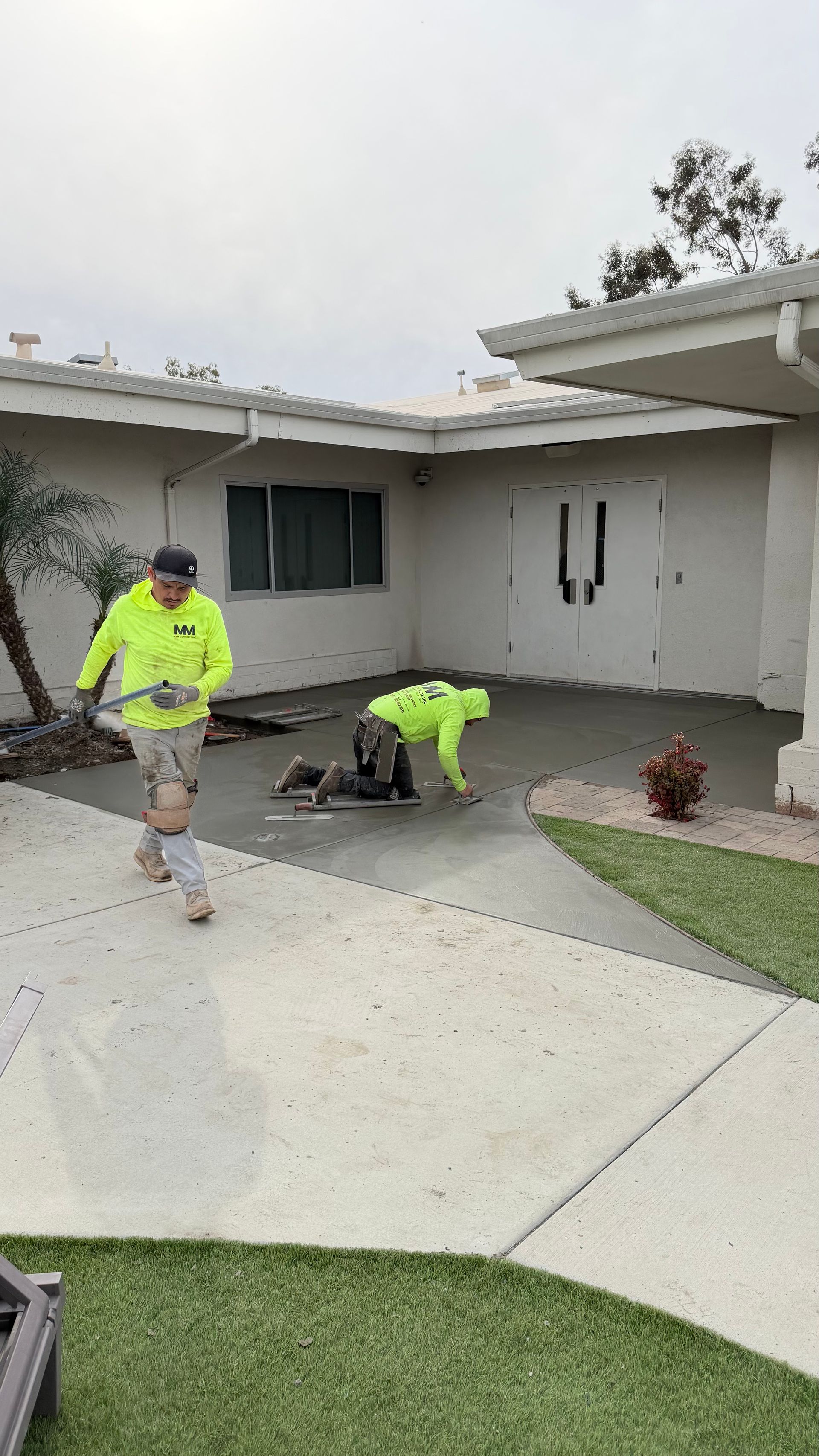 Two workers in neon shirts repairing a concrete walkway outside a house