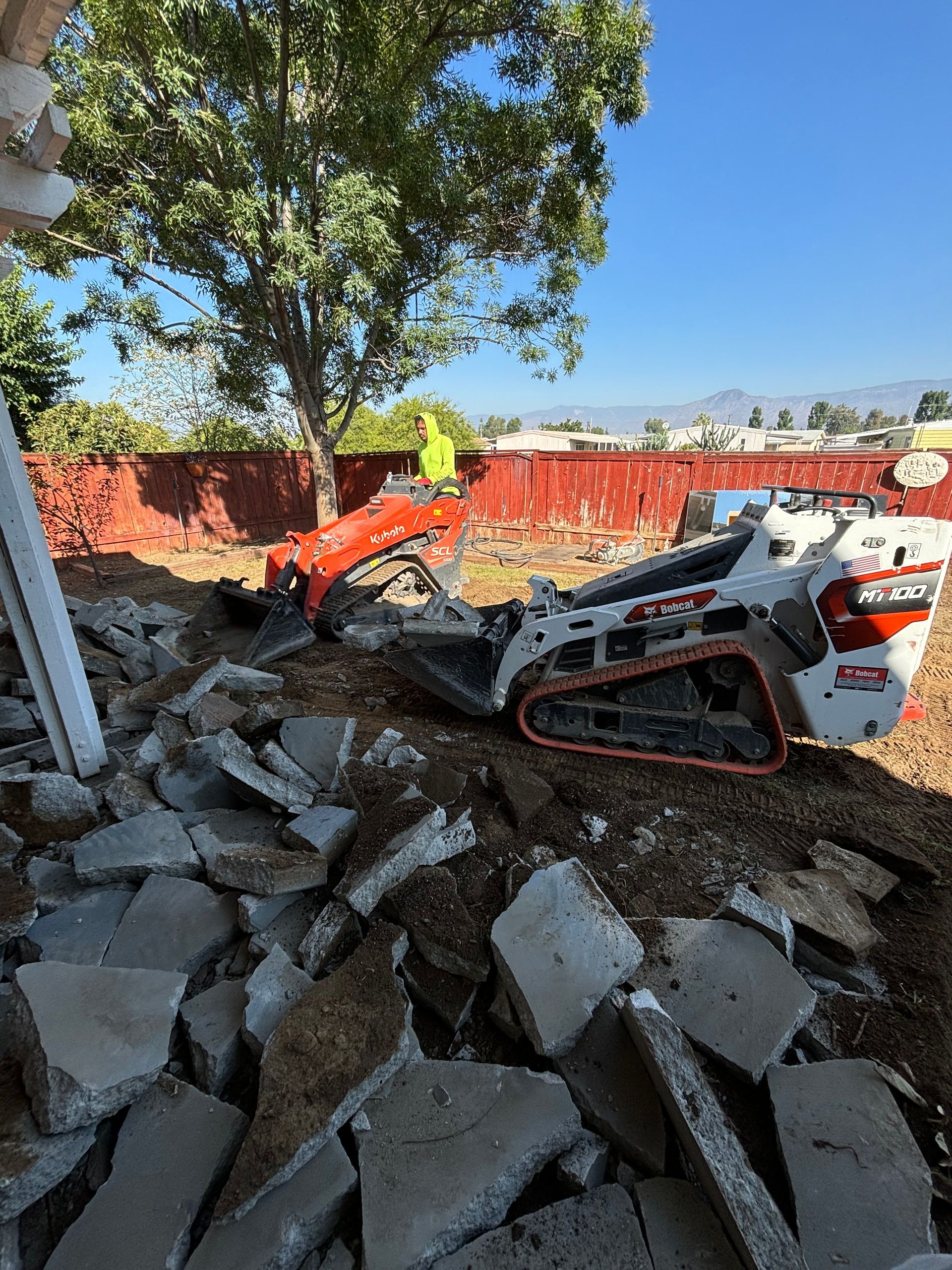 Worker operates a compact excavator amid broken concrete rubble at a construction site.