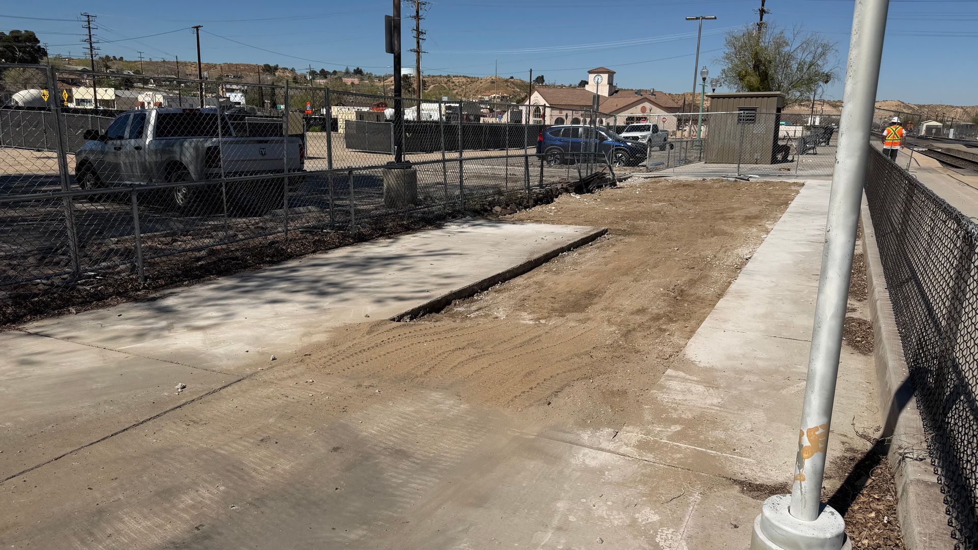 Dirt path beside a fenced sidewalk under construction on a sunny street.