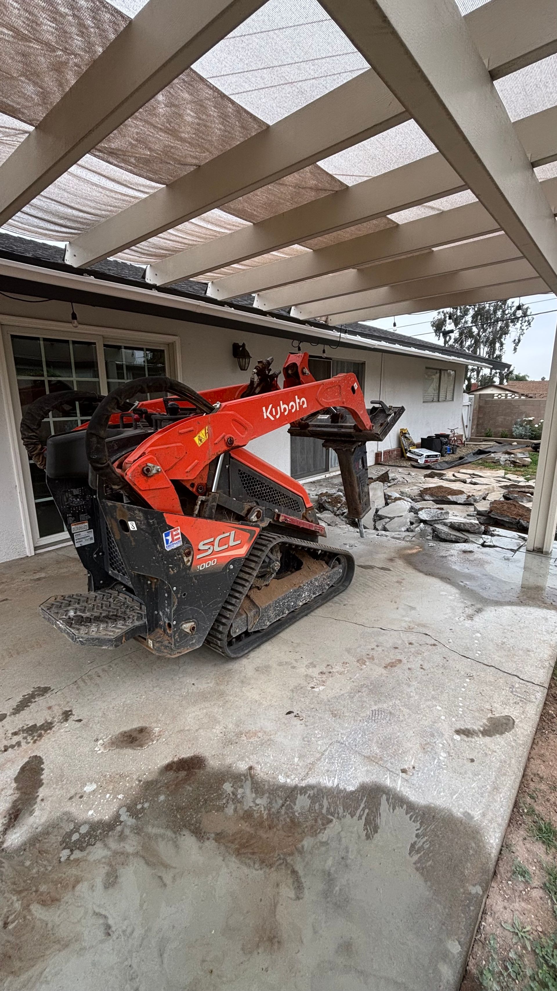 Orange tracked excavator in a damaged, debris-strewn building interior under a broken roof.