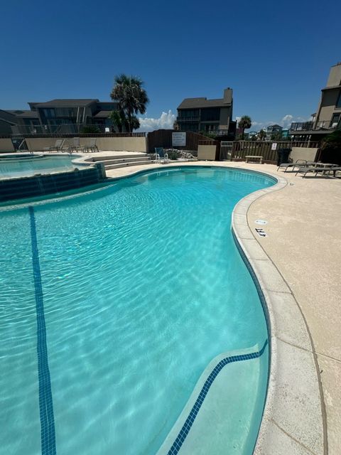 Swimming pool with clear blue water under a bright blue sky. Buildings and lounge chairs are visible.