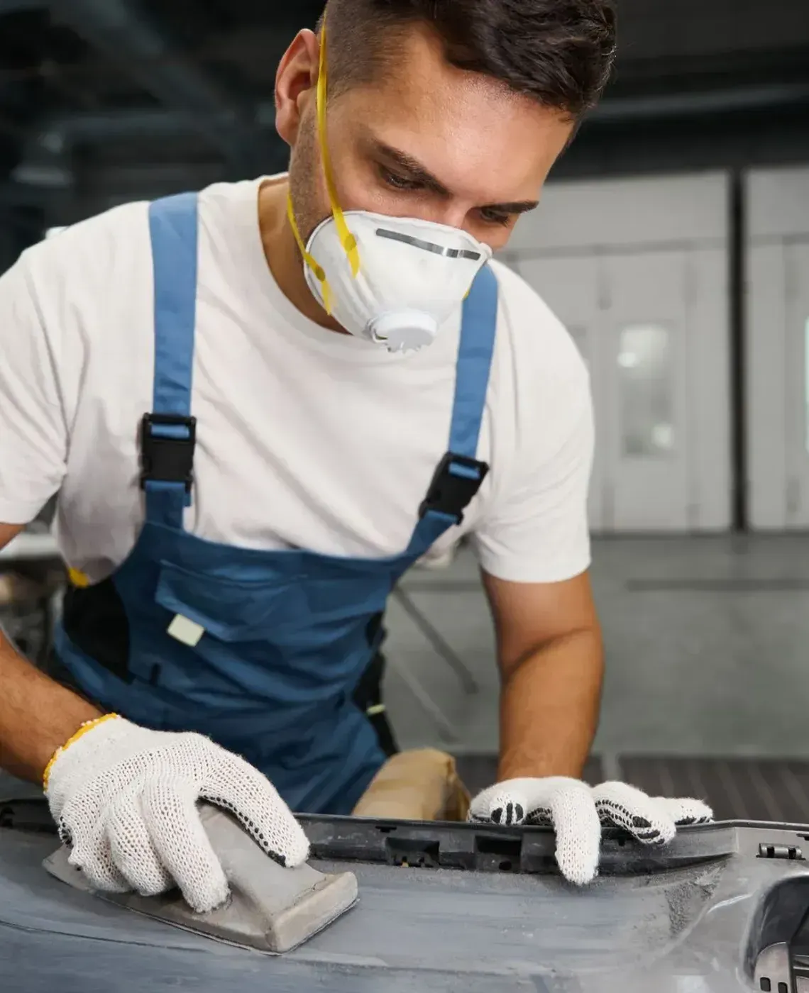 Mechanic in blue overalls and respirator sanding a car part in a workshop