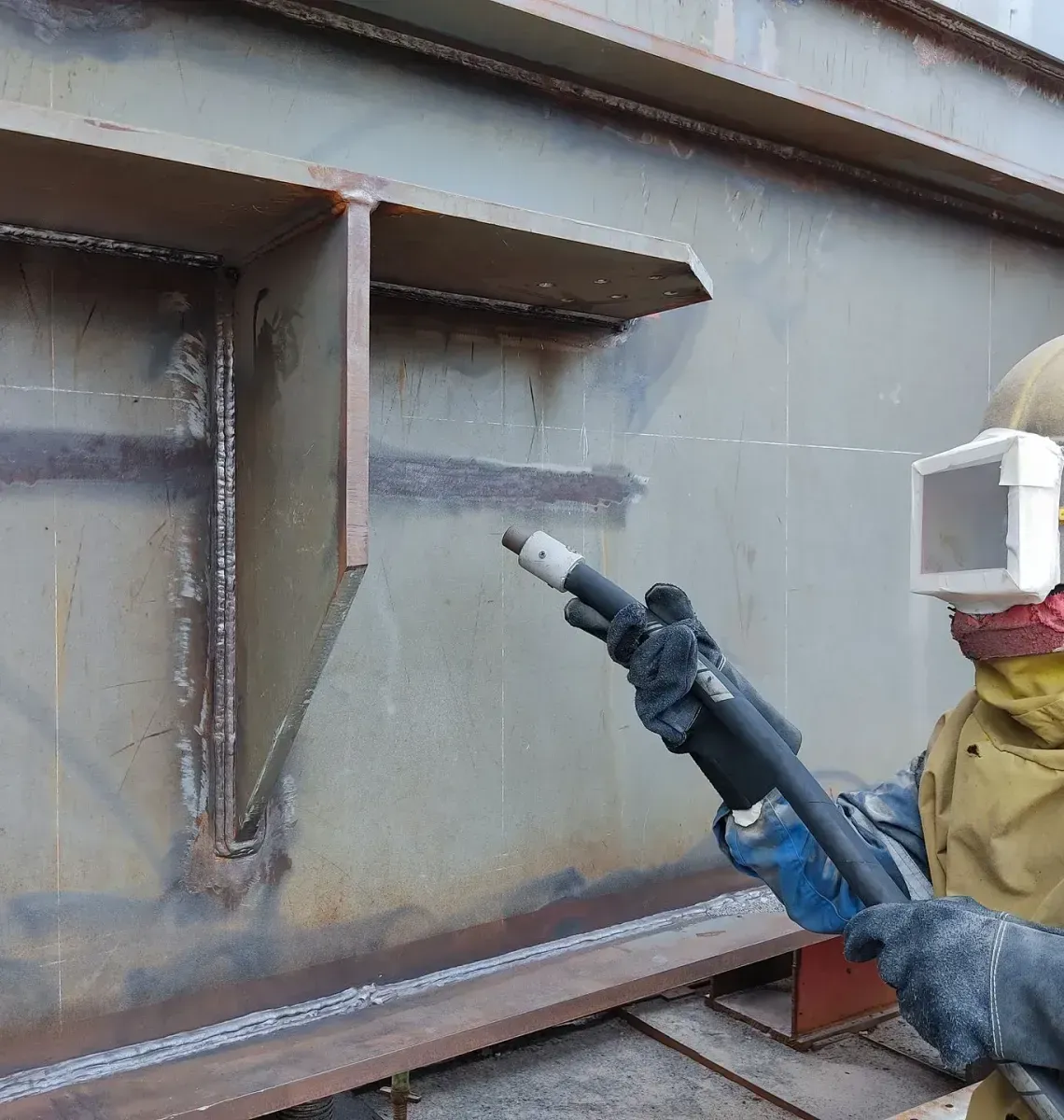 Worker in protective gear using a blue spray gun to coat a gray metal structure.