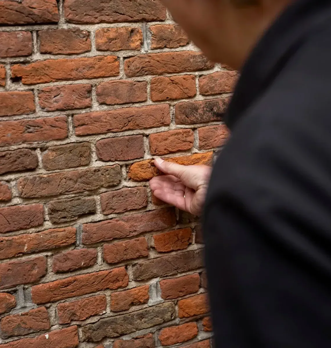 Hand placing a brick on a brick wall with mortar, against a red brick background