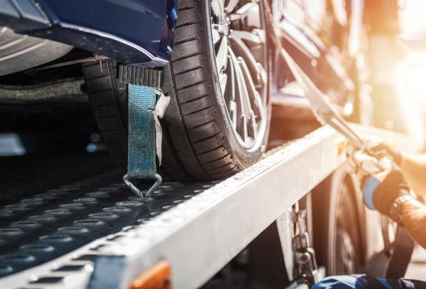 A car being secured to a flatbed tow truck with blue straps, sunny outdoors.
