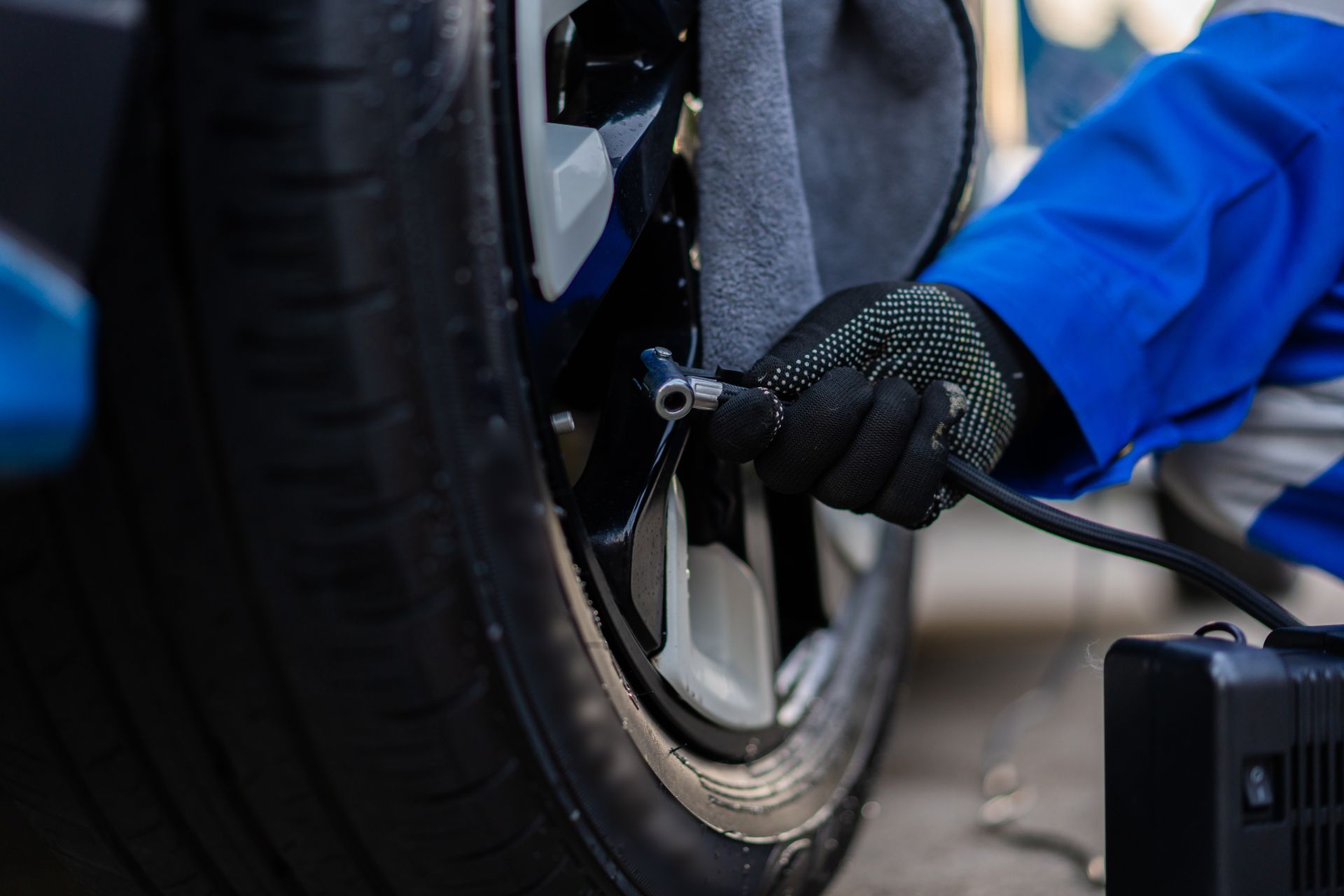 A person inflating a car tire with a black air compressor. They wear work gloves and a blue jacket.