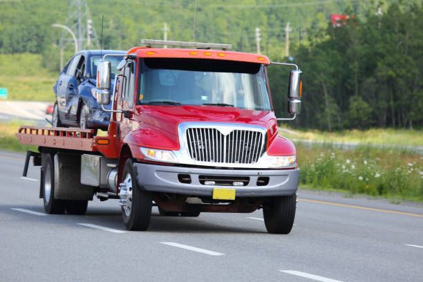 Red tow truck carrying a damaged blue car on a highway.