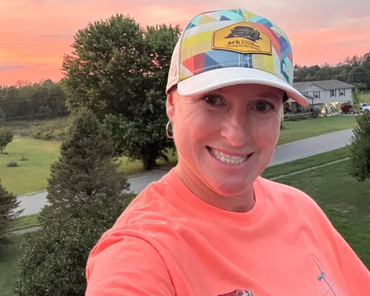 Smiling person in a coral shirt and colorful cap outdoors at sunset with trees and houses behind them
