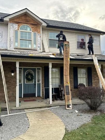 Workers on ladders installing a wooden support beam on a two-story house porch.