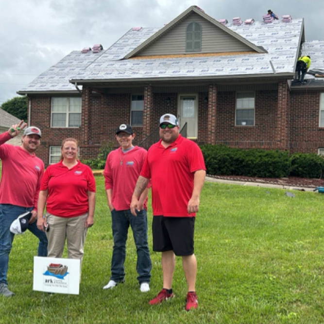 Volunteers in red shirts pose on a lawn outside a brick house with a roof crew working behind them