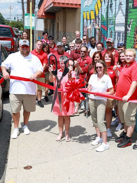 People at a ribbon-cutting ceremony on a sunny street, with a large red ribbon and bow.