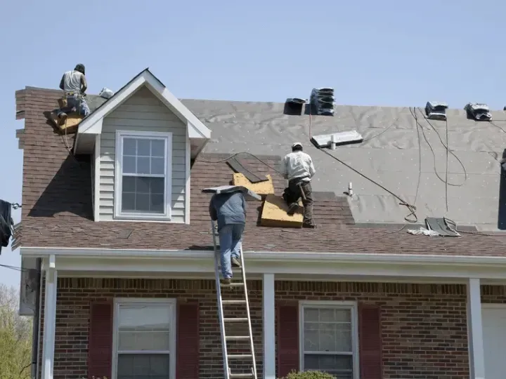 Workers reroofing a brick house, with one man on a ladder and shingles on the roof.