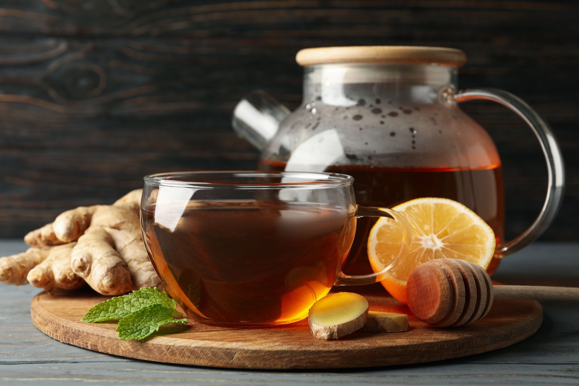 A cup of hot tea on a wooden board with ginger, lemon slices, mint, and a honey dipper. A cup of hot tea on a wooden board with ginger, lemon slices, mint, and a honey dipper.