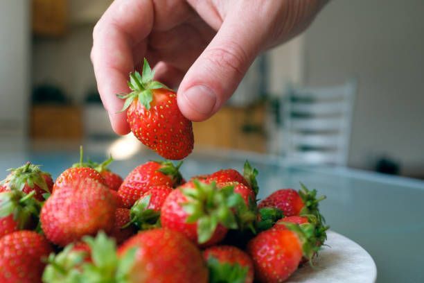 Hand reaching for a ripe strawberry from a plate of strawberries. Bright red berries, white plate.