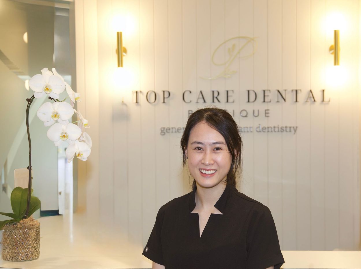 Smiling person standing in a dental clinic lobby beside white orchids and a “Top Care Dental” sign