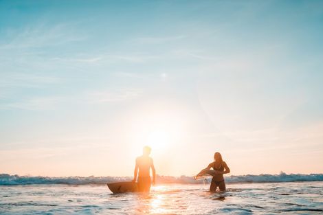 Two people wading in the ocean at sunset, silhouetted against a bright sky.