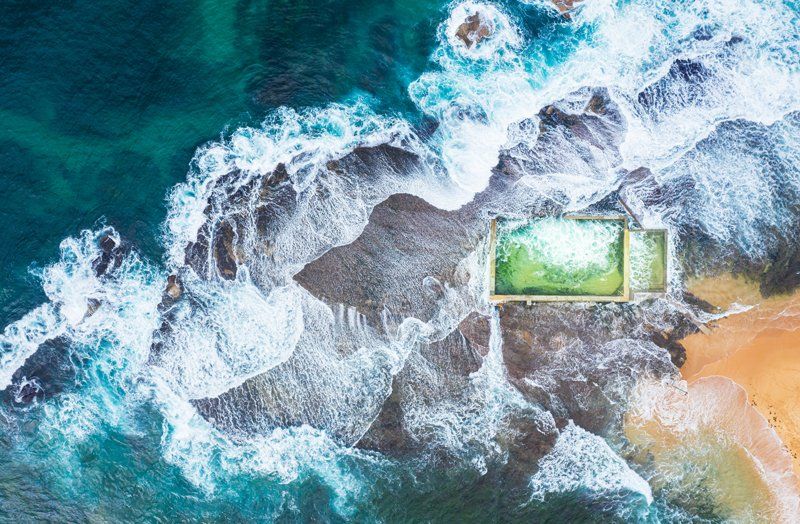 Aerial view of ocean waves crashing on a rocky beach beside a small green-roofed building.