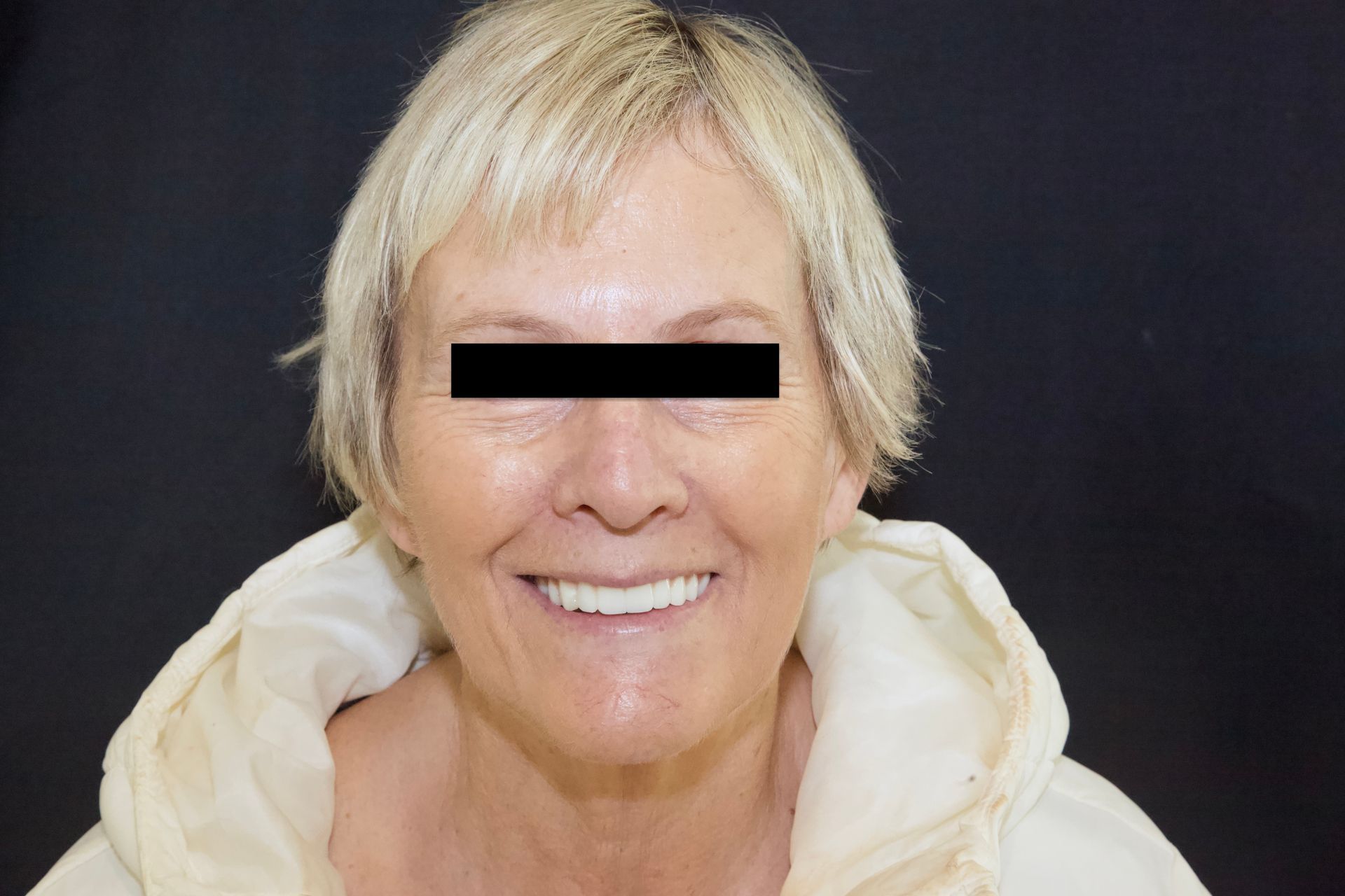 Smiling person with blonde hair against a dark background, wearing a light hoodie