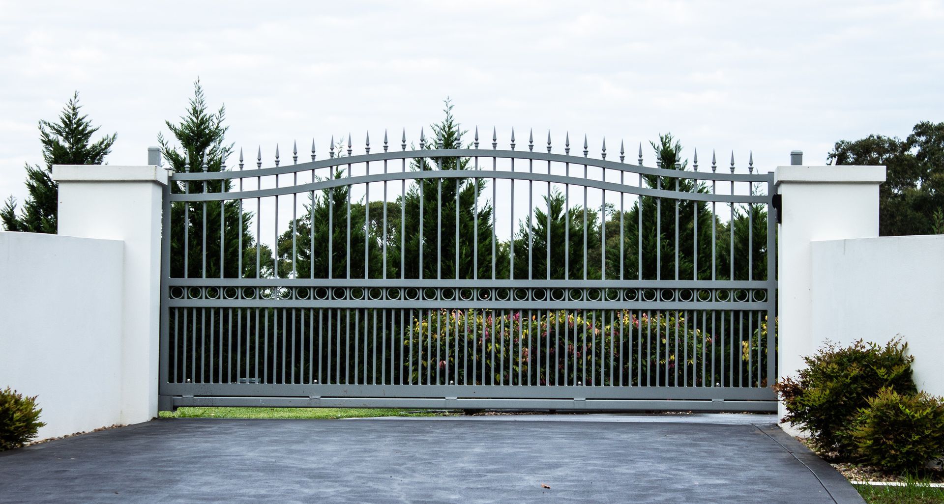 Grey wrought iron driveway gates set in a white brick fence with trees behind