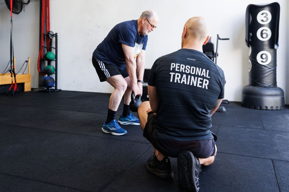 Man lifting kettlebell watched by personal trainer in a gym.