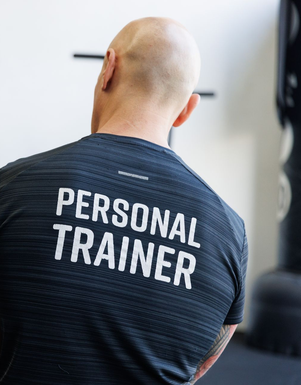 Personal trainer observes a man lifting kettlebells in a gym. The trainer's shirt reads 