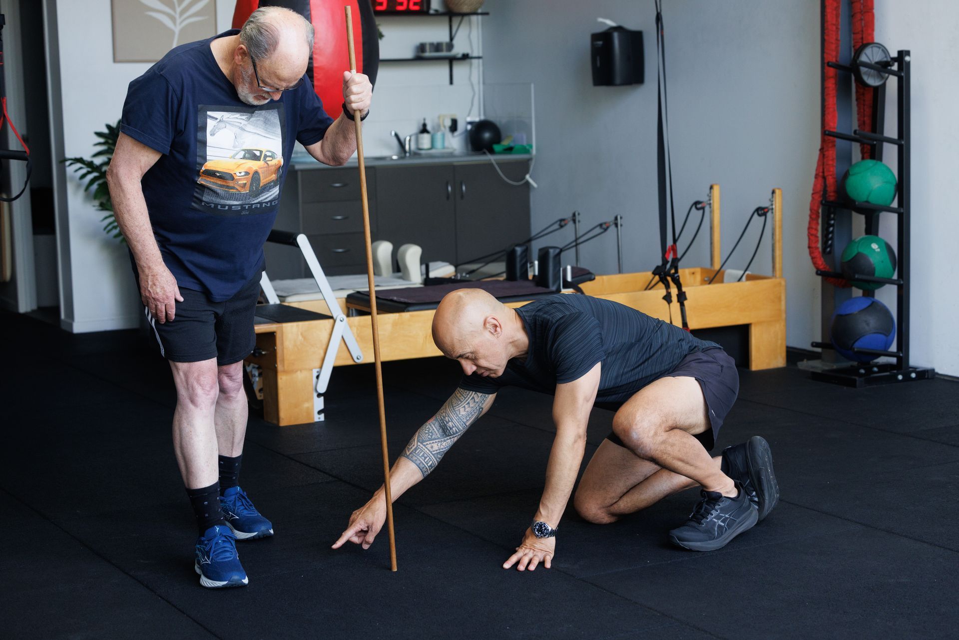 Man in shorts, using a stick with trainer for exercise in a gym.