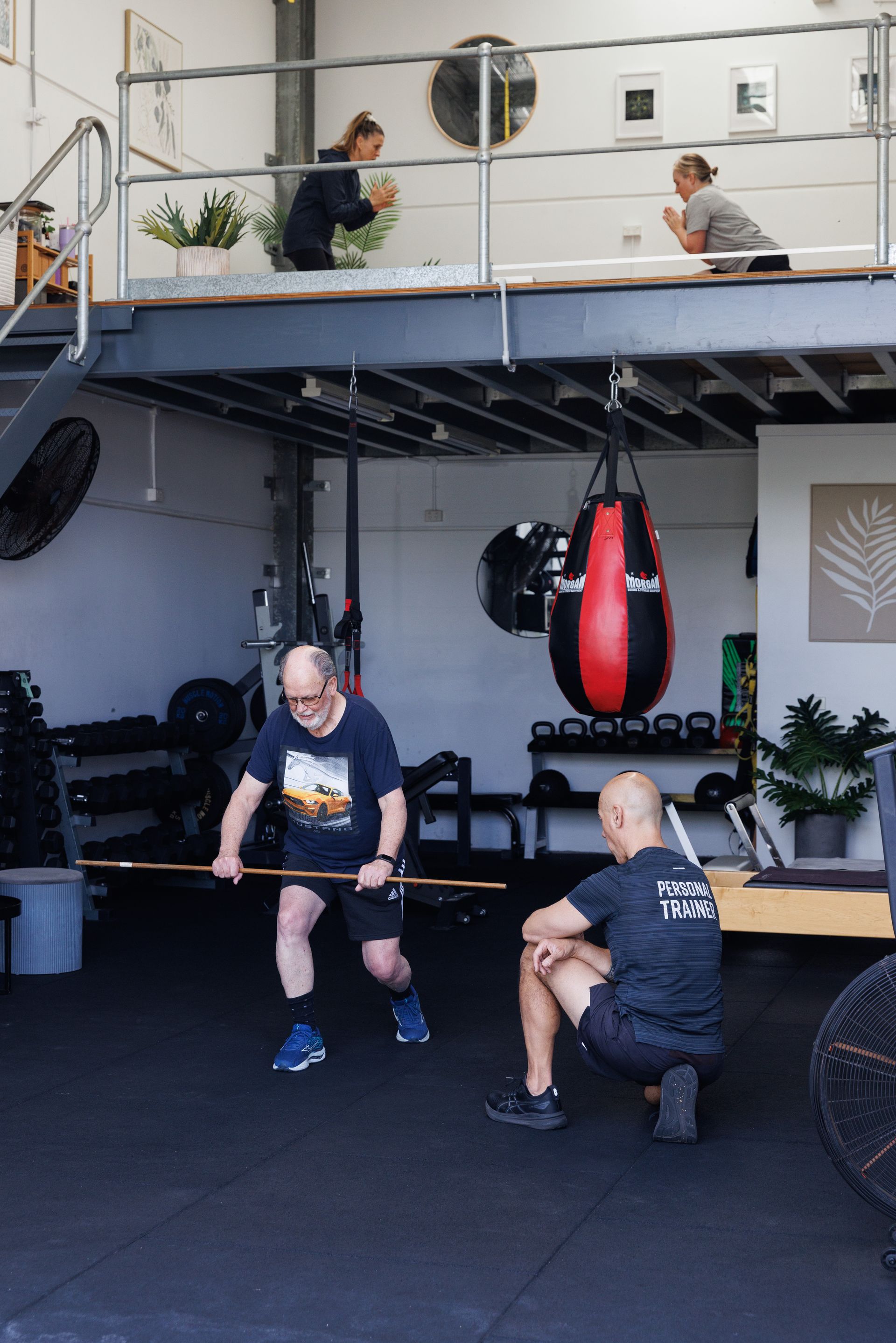 A man exercises with a trainer in a gym. A yoga warmup on a loft above.