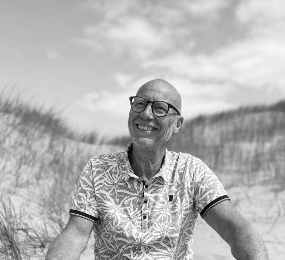 Een lachende kale man met een bril zit op een zandstrand met hoog gras en een bewolkte hemel.