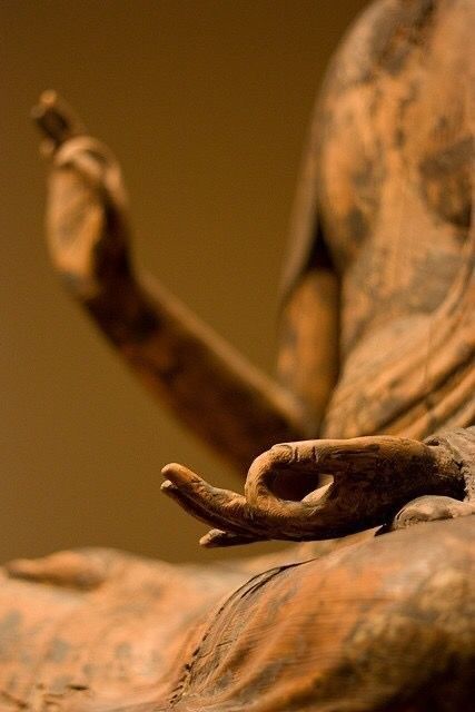 Close-up van de hand van een bronzen beeld in een meditatiehouding tegen een warme bruine achtergrond.