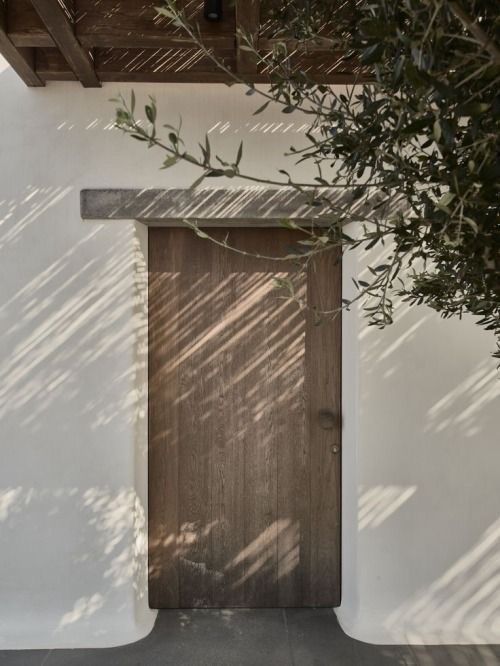 Witte stucmuur met een houten deur, waar de schaduwen van bomen doorheen schijnen en die omlijst wordt door olijftakken.