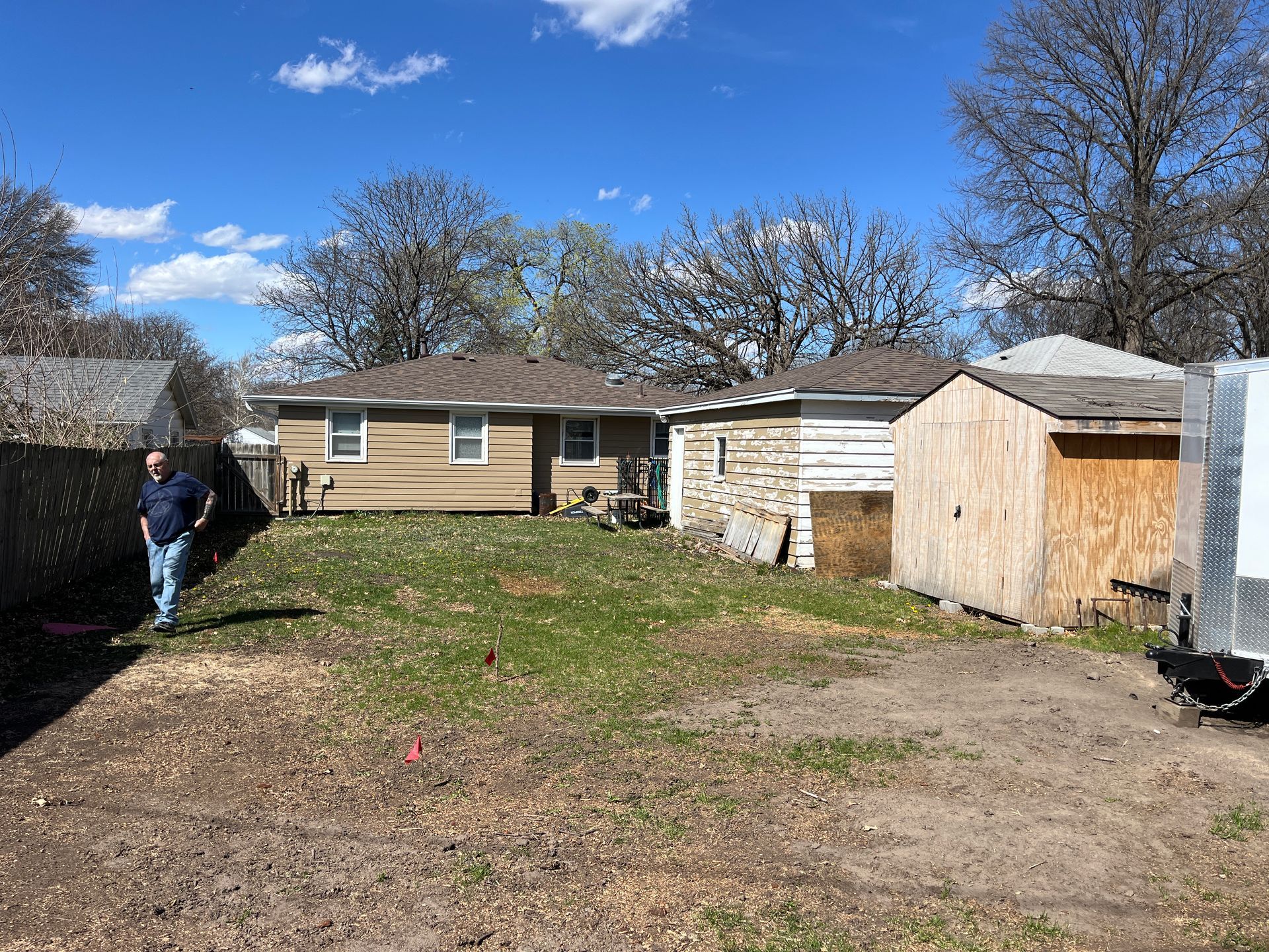 A man stands in a small, mostly-bare backyard with a house, shed, and trailer. Bright blue sky overhead.
