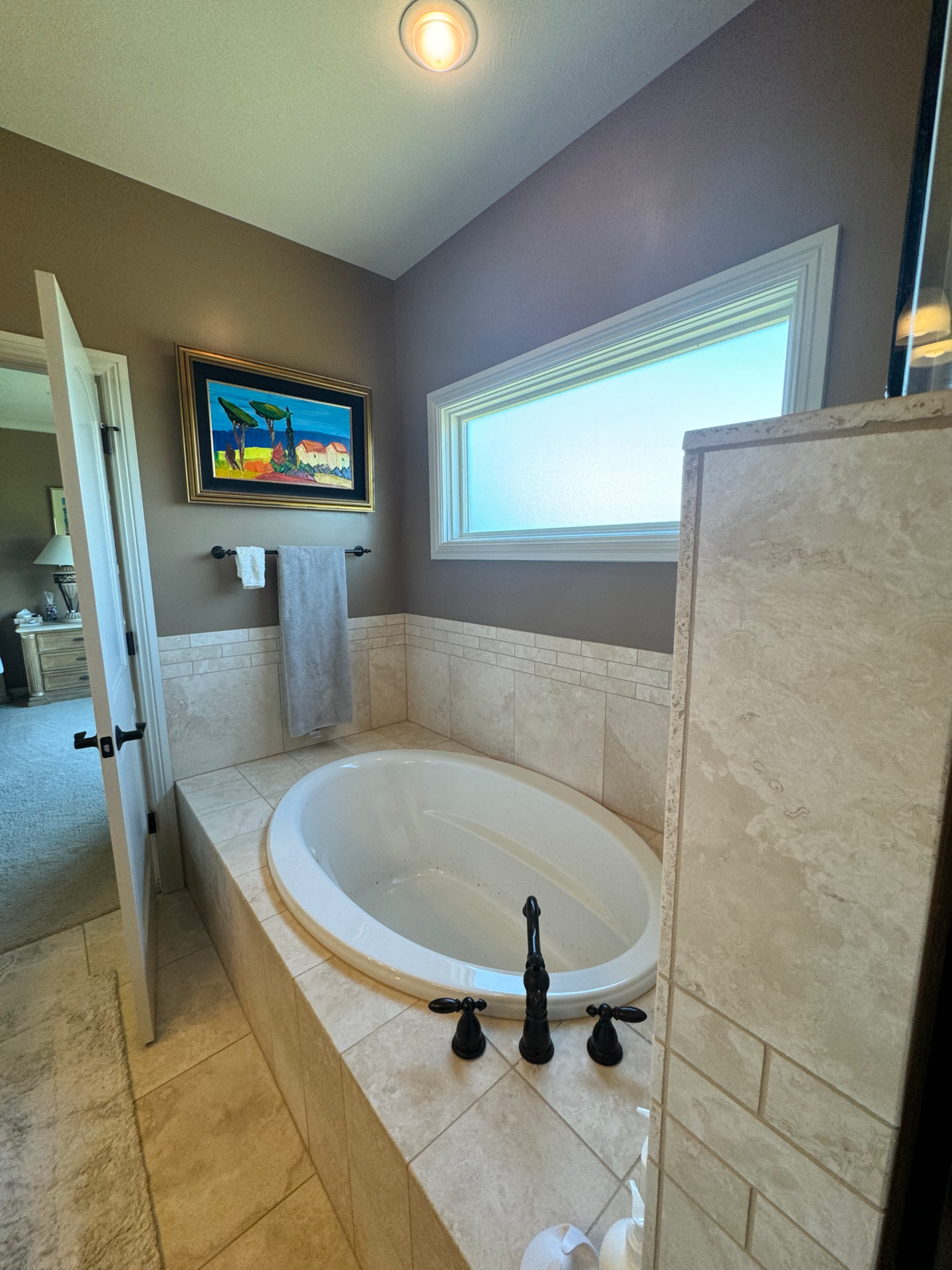 Bathroom with oval bathtub, neutral-toned tile, painting above the tub, and a window.