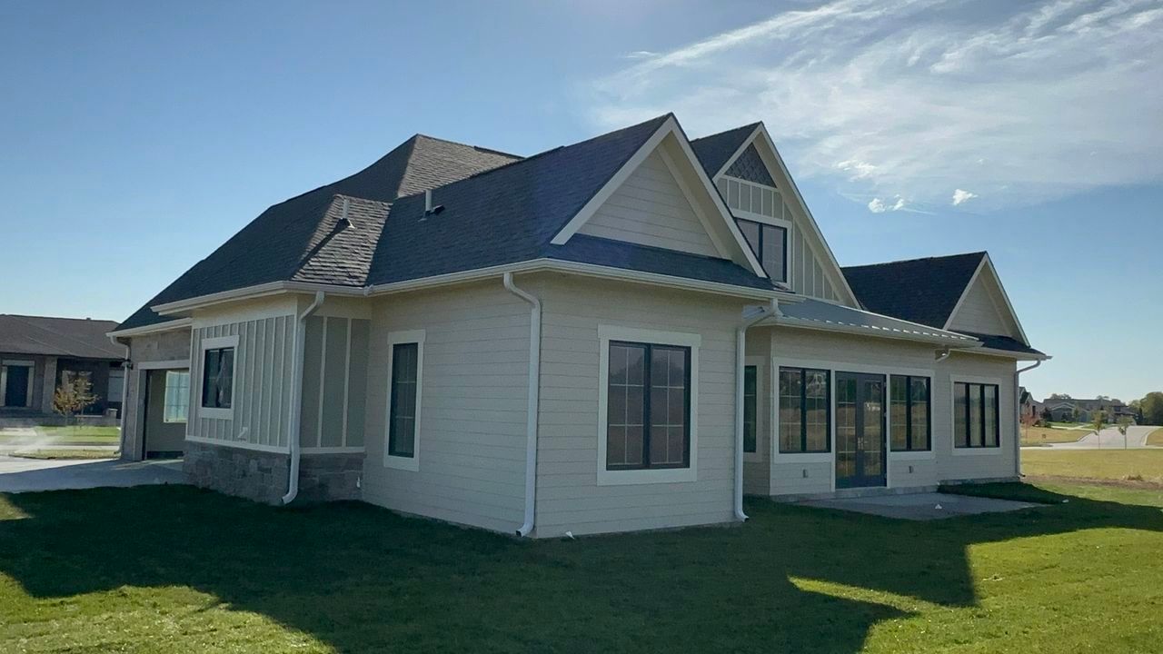 The side and rear of a new home with beige lap and board-and-batten siding, a stone base, white gutters, dark-framed windows, French doors and fresh sod.