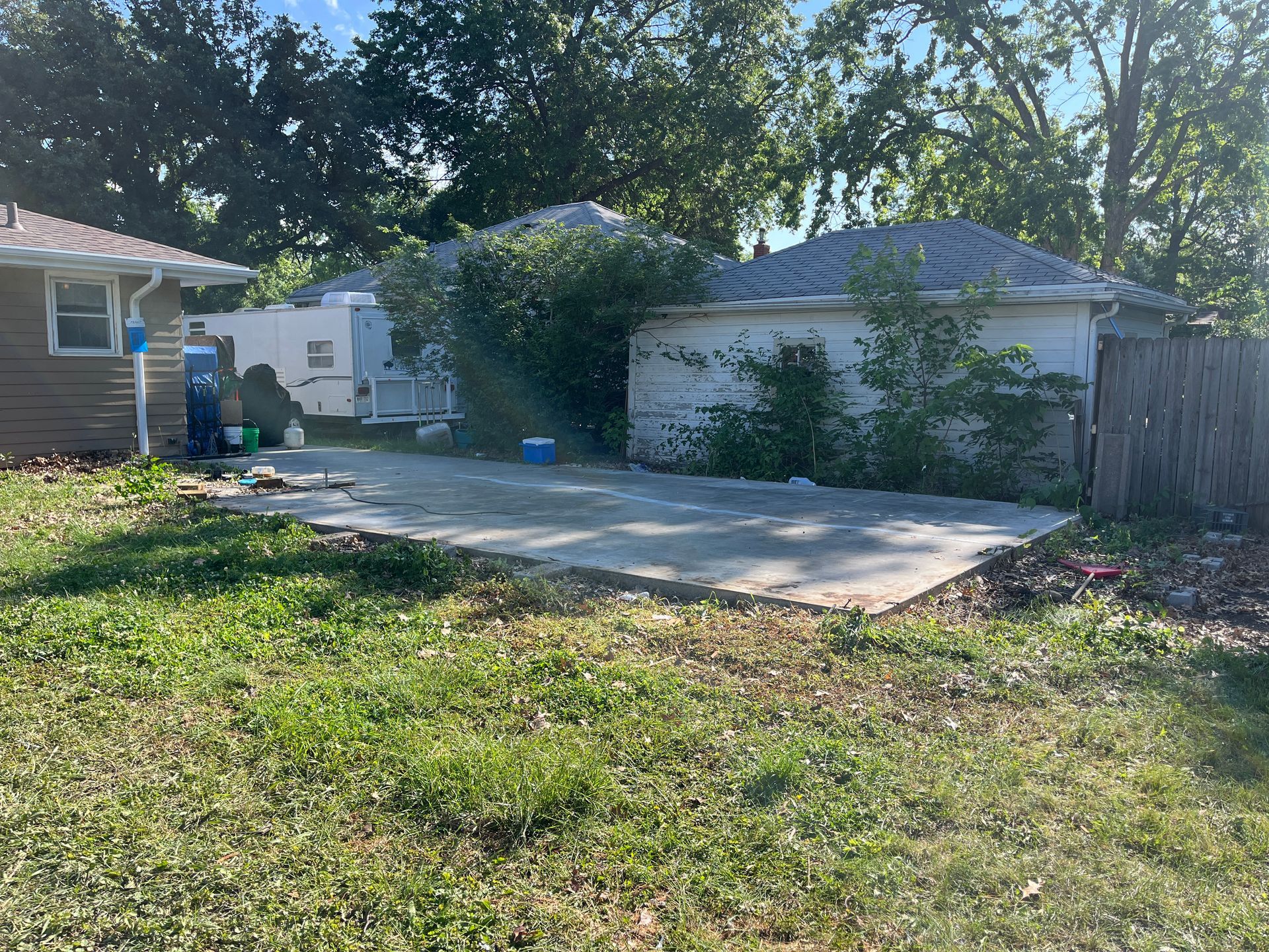 Backyard view with a concrete slab, small garage, and overgrown greenery under a sunny sky.