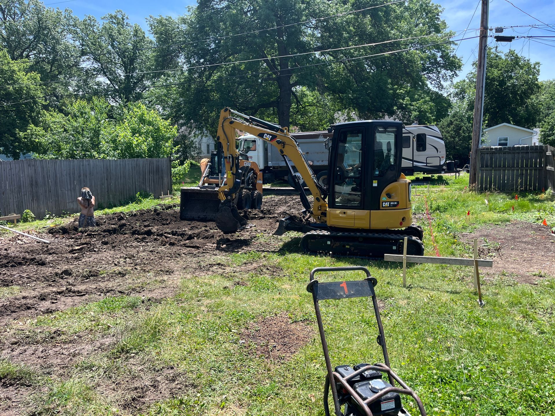 A yellow excavator digs into a dirt patch in a yard on a sunny day. A lawnmower sits nearby.