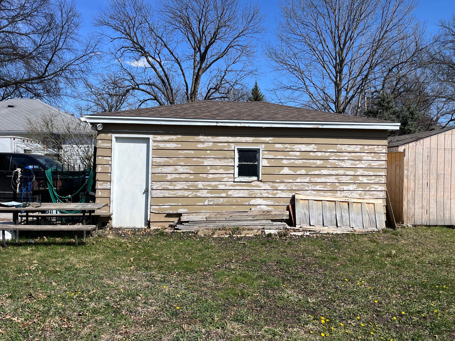 A weathered, beige shed with peeling paint, a white door, and small window sits in a grassy backyard on a sunny day.