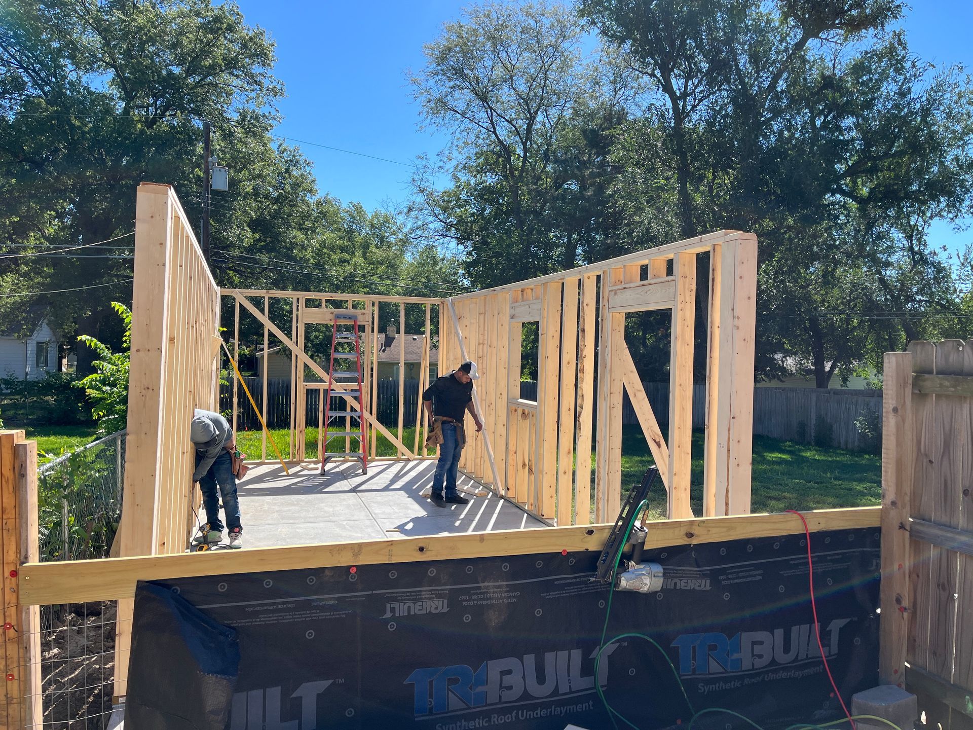 Two workers building a wooden structure outdoors; one on the wall, another standing near a window frame.