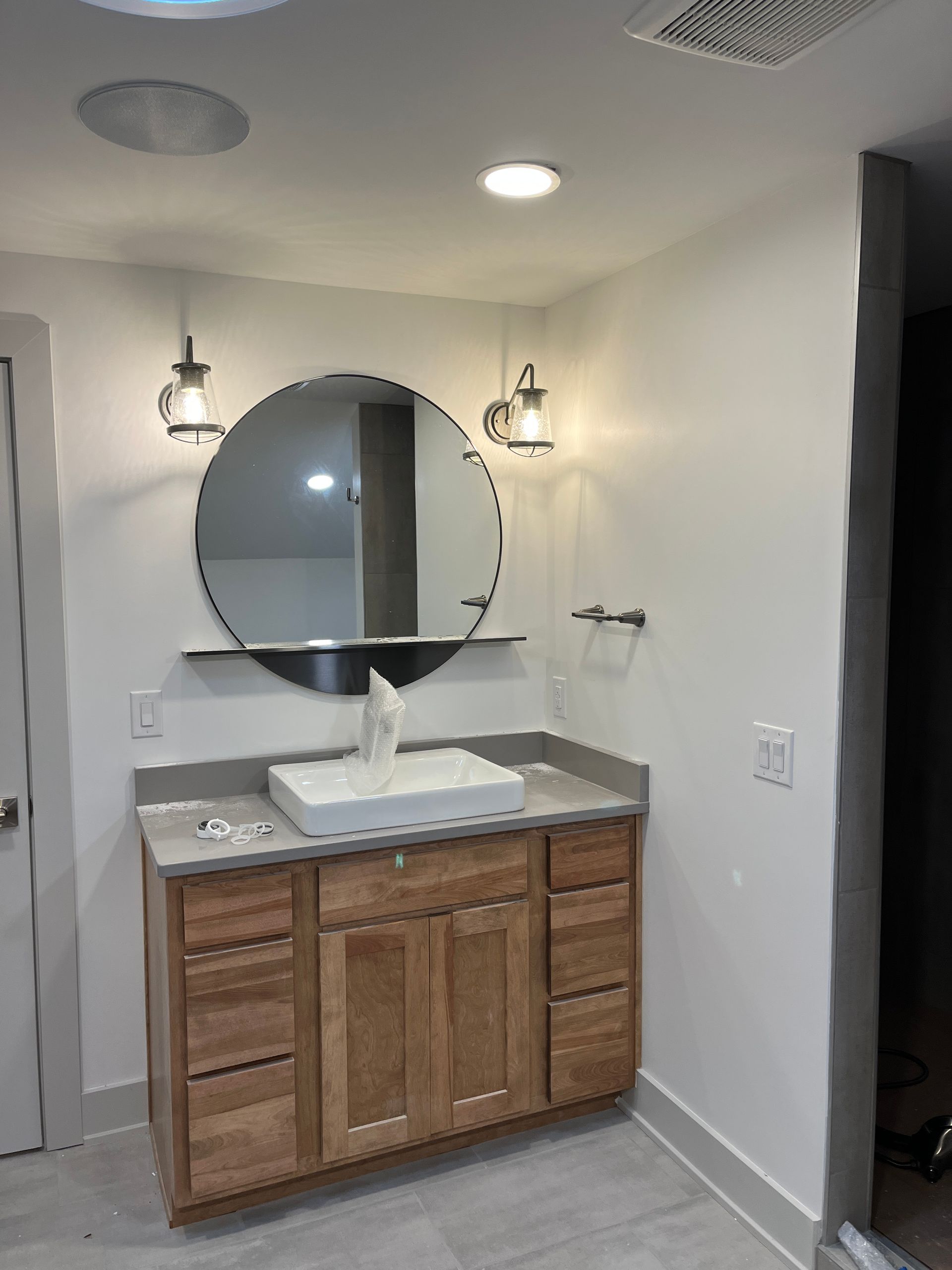 A bathroom with a wooden vanity, round mirror, and sconce lighting. White walls, a white sink, and light gray countertop.