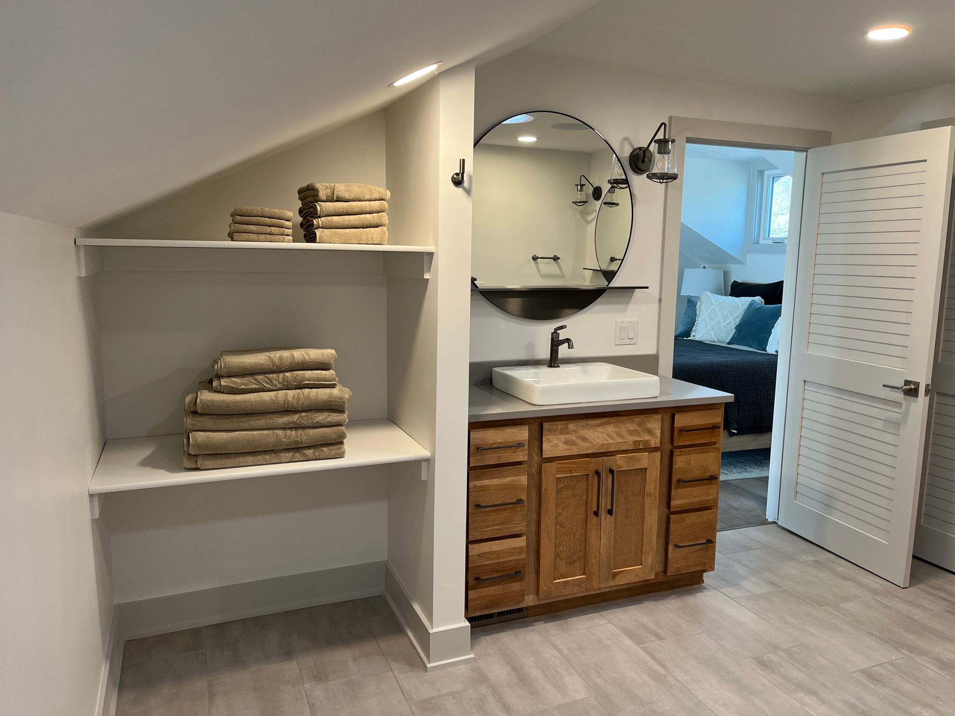 Bathroom with open shelving, wood vanity, round mirror, and doorway to bedroom. Light walls, flooring, and towels.