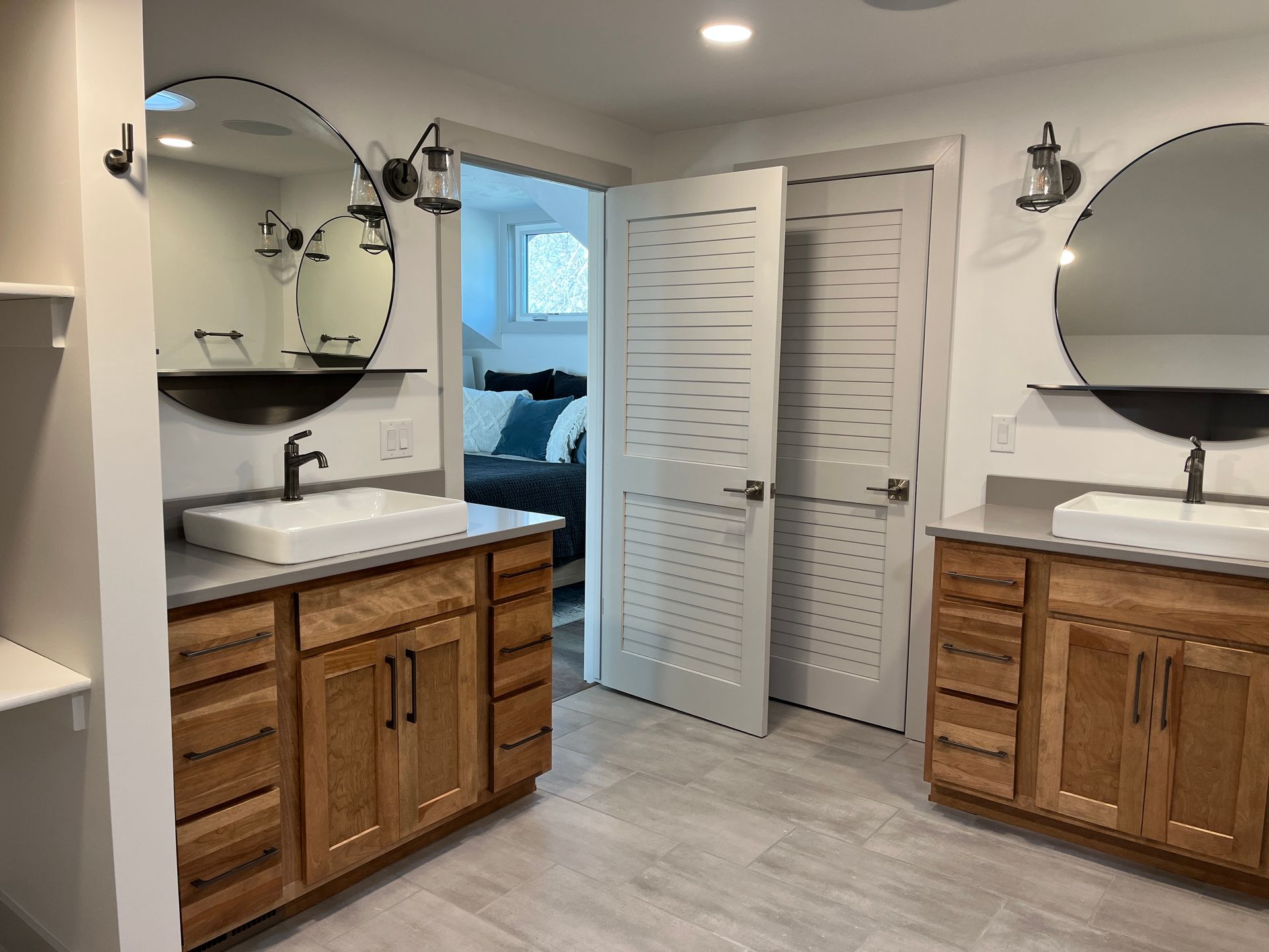 Bathroom with two wood vanities, round mirrors, and white sinks, viewed from the doorway leading to a bedroom.