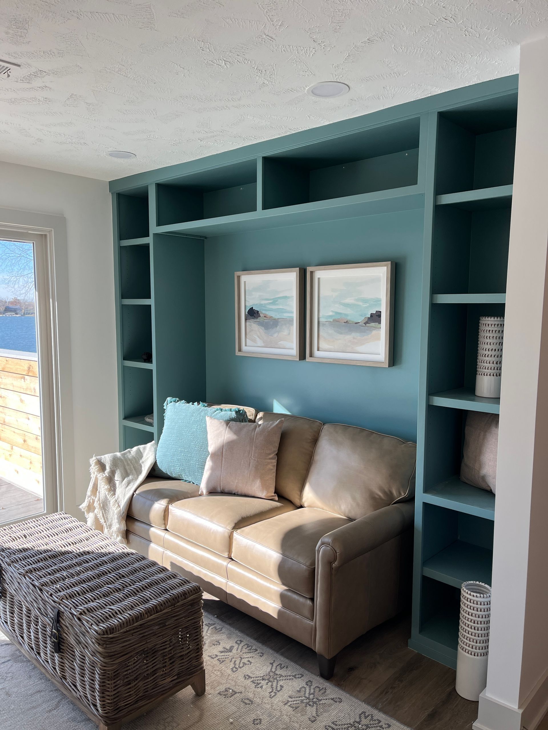 Built-in teal shelves frame a leather sofa with pillows. Artwork hangs above, and a woven chest sits nearby.