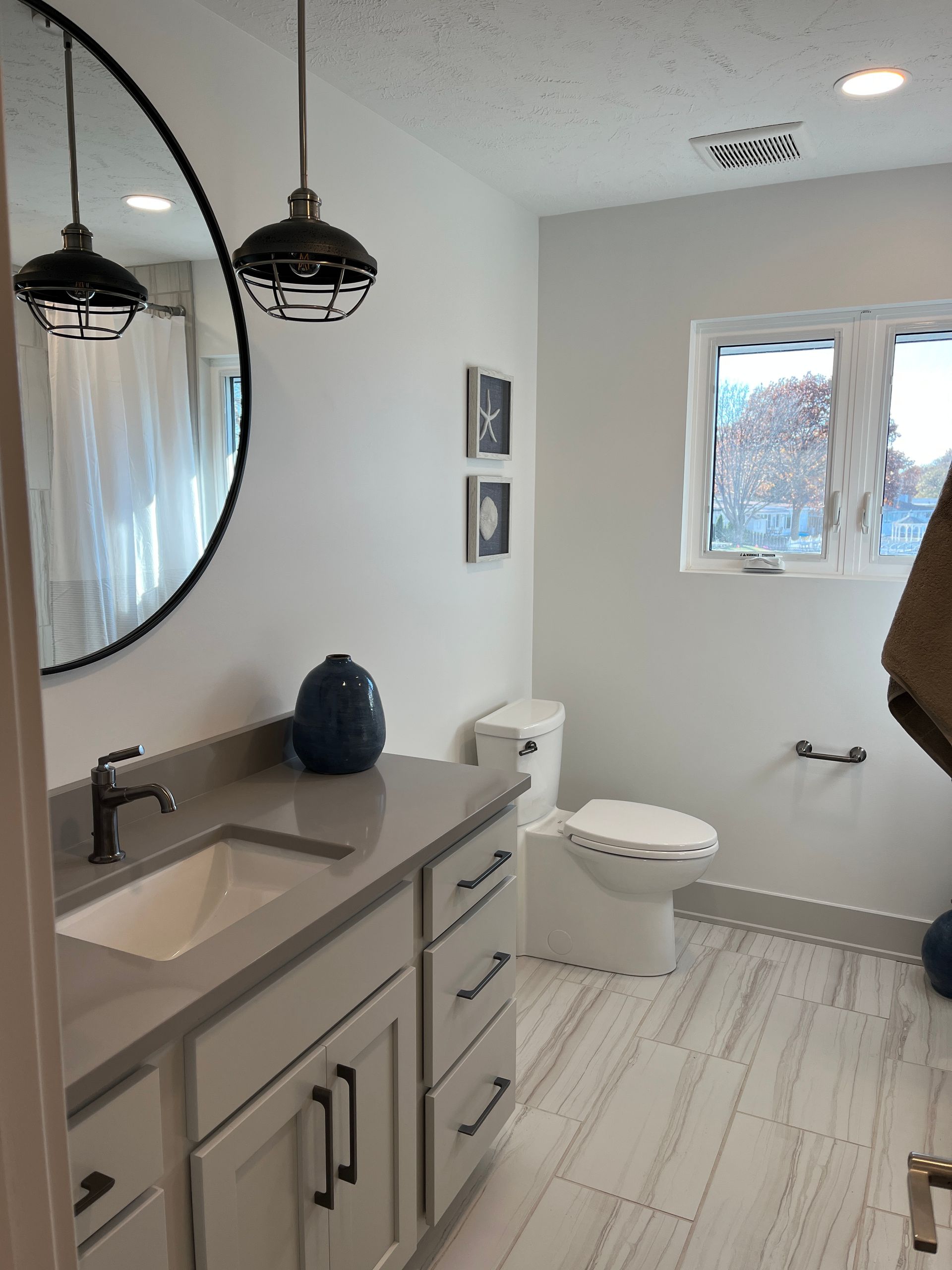 Modern white bathroom with gray vanity, round black mirror, and two pendant lights.